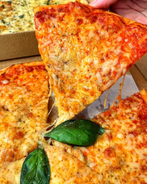 Freshly baked cheese pizza slice with basil leaves being pulled from the pie